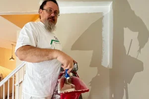 A man wearing glasses and a white t-shirt, working with Tribble Painting, uses Sherwin-Williams paint to create an accent wall with a brush. Standing near a staircase in a Southeast Michigan home—such as one in Ann Arbor, Ypsilanti, or Novi—he holds a red paint tray while his shadow is cast on the freshly painted wall. by Tribble Painting